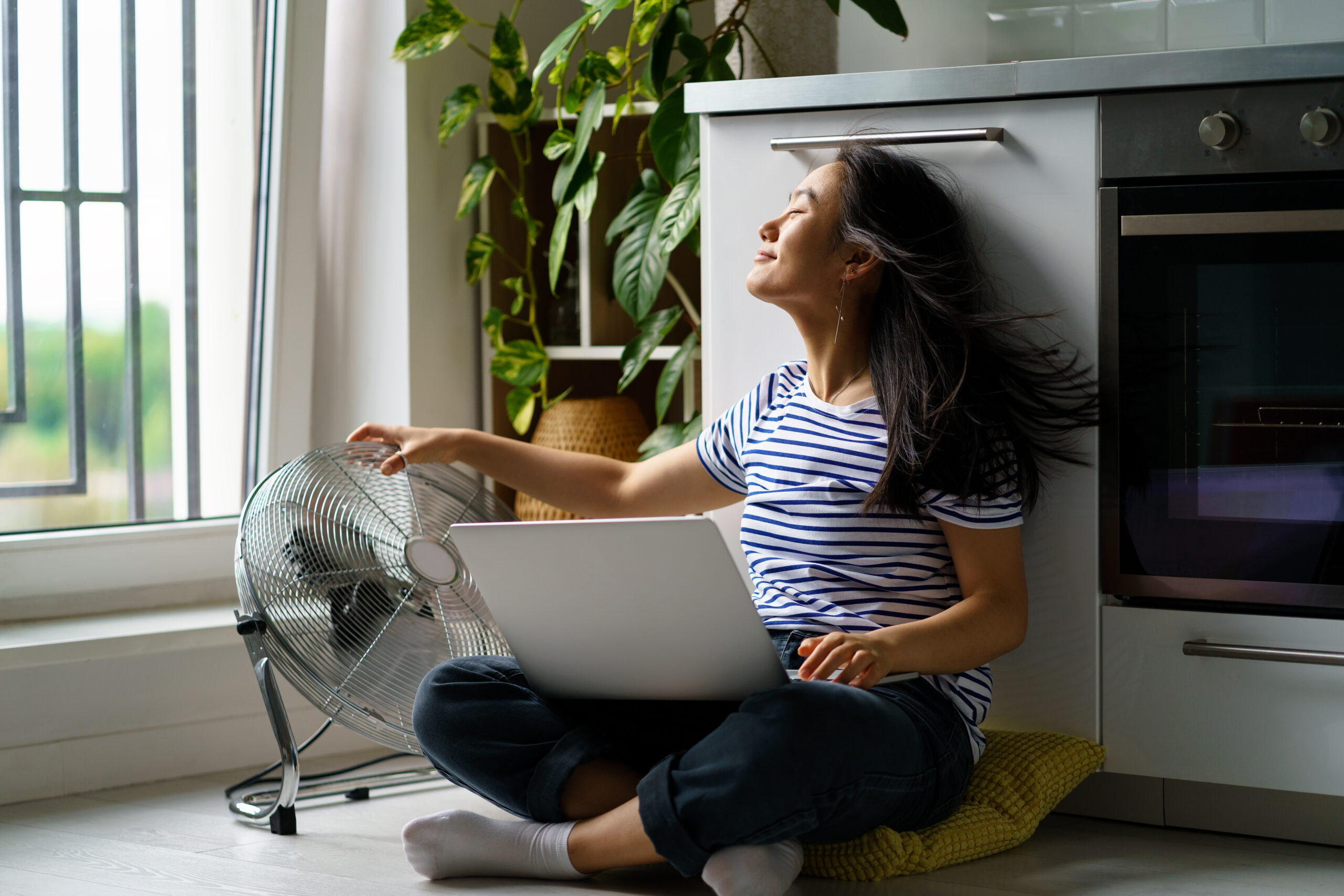 Satisfied Asian woman sits on floor with laptop on crossed knees, enjoying wind from electric fan