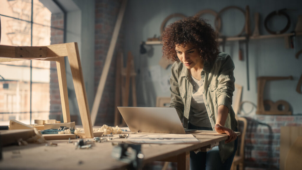 Woodworker Using Laptop Computer and Building a Wooden Chair. Black Multiethnic Female Carpenter Working in a Studio in Loft Space with Tools on the Walls.