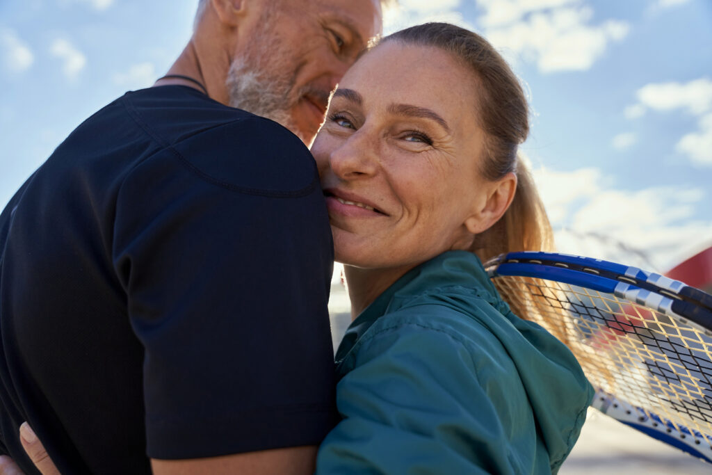 Portrait of active mature couple looking happy while embracing each other outdoors, ready for morning workout on tennis court. Sport, healthy lifestyle concept