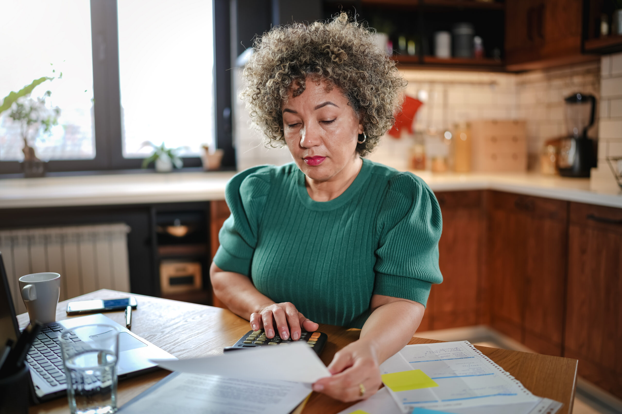 A mature woman with curly gray hair works on a laptop in a cozy home kitchen. The setting is relaxed, with natural light coming in through the window, creating a productive atmosphere