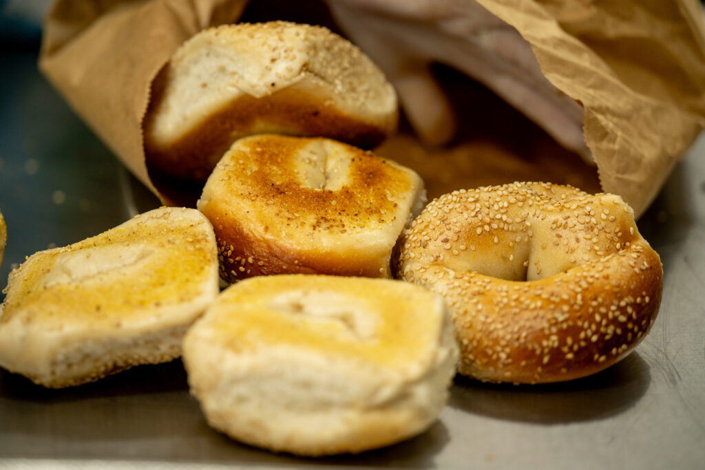 Fresh sesame and plain bagels spill from a brown paper bakery bag onto a metal kitchen counter. A takeout and bakery purchase scene showing plentiful food ready to eat.