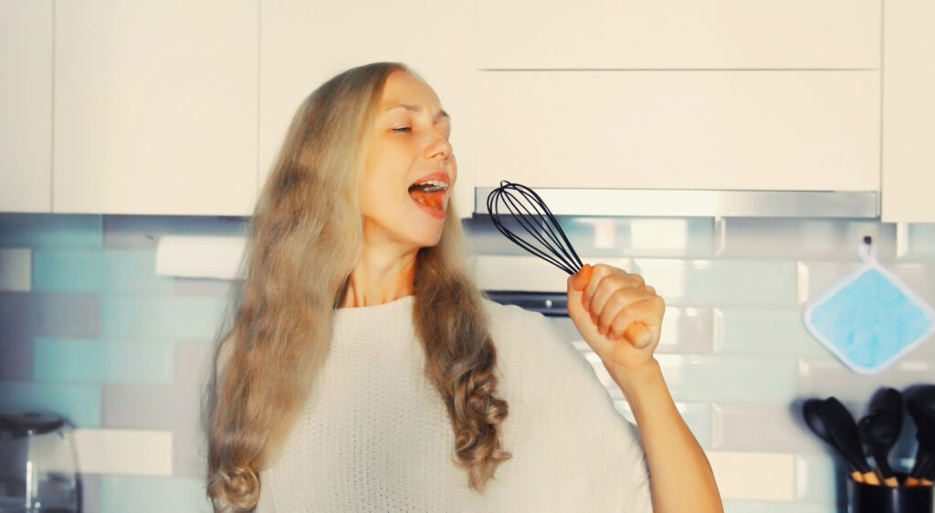 Happy cheerful middle aged woman singing with whisk having fun in the kitchen at home